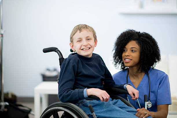 A nurse is with a child in the hospital that has cerebral palsy. The little boy is sitting in a wheelchair. He is smiling and looking at the camera.